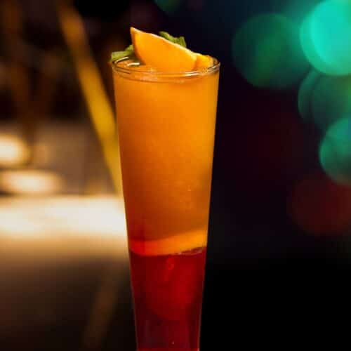 A layered Tequila Sunrise cocktail made with Casa México Tequila in a tall glass, showing a vibrant red-to-yellow gradient over ice, garnished with an orange slice.